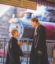 Two children wearing wizard robes queue in front of the Hogwarts Express at Hogsmeade Station.